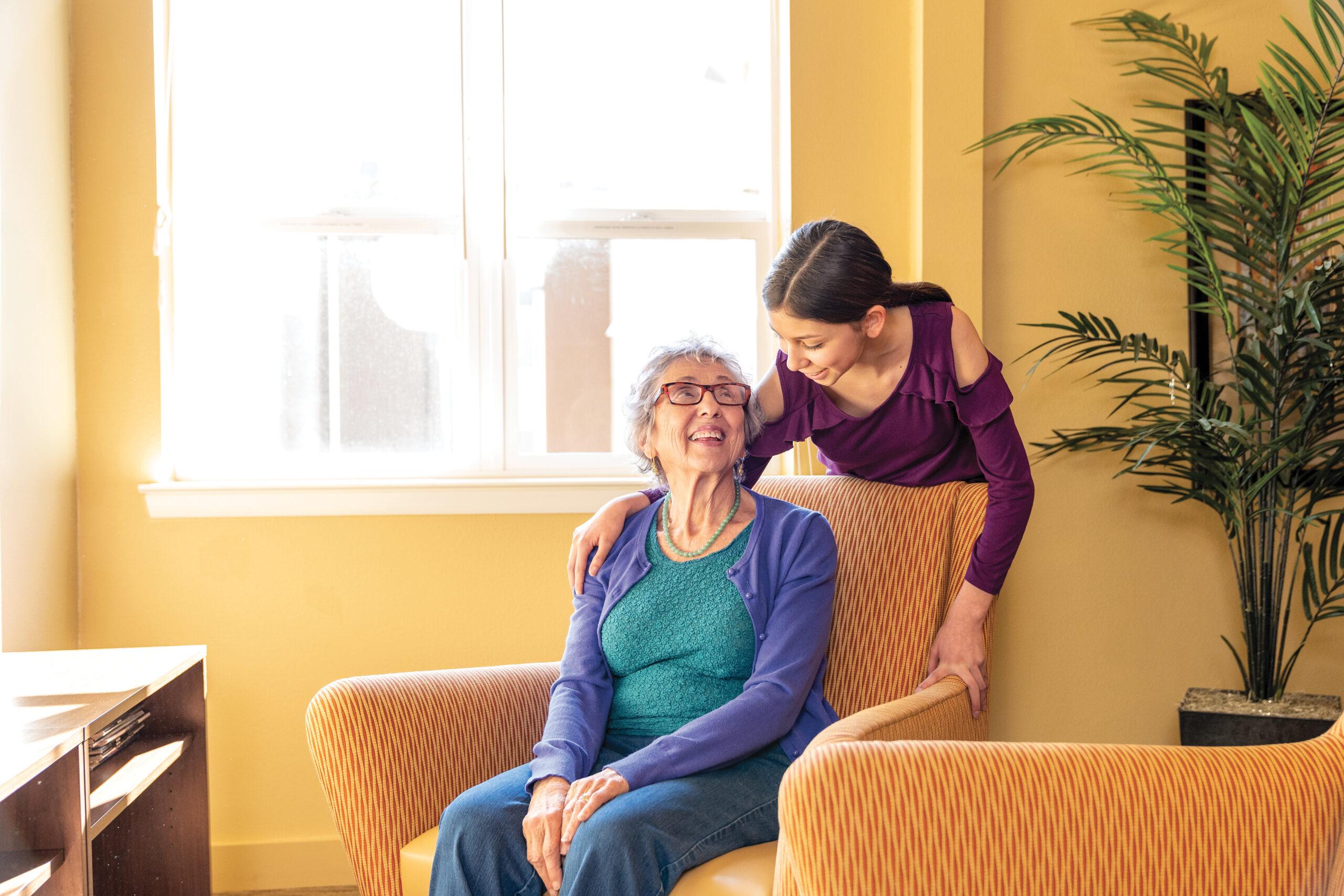 A young girl leaning over an elderly woman seated in an armchair, both smiling warmly.