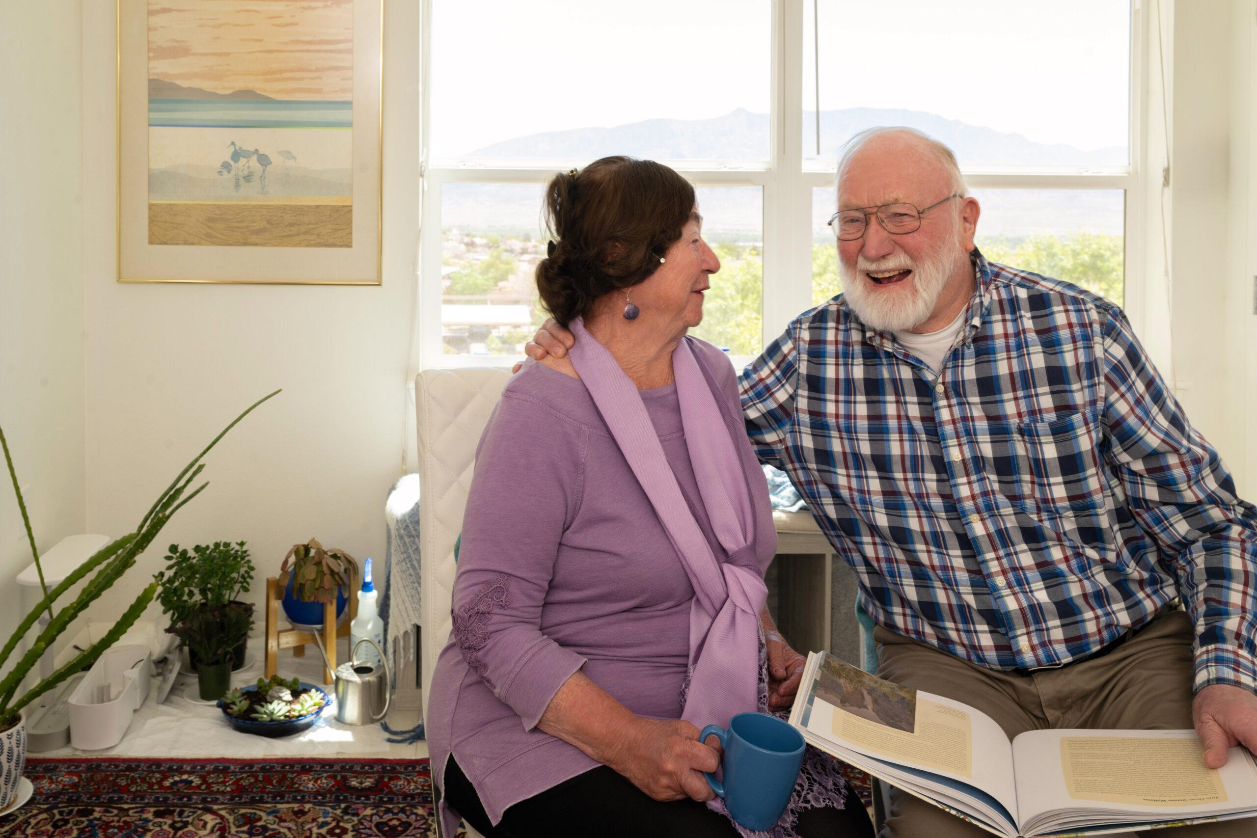 An older man and woman sitting side by side on a sofa next to a window, laughing warmly while looking at each other.