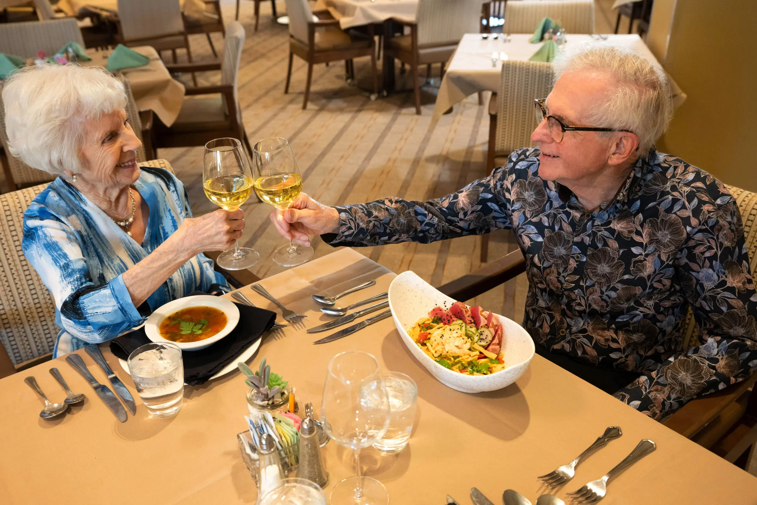 An elderly couple clinking glasses of white wine over a restaurant table set with soup and a salad bowl.