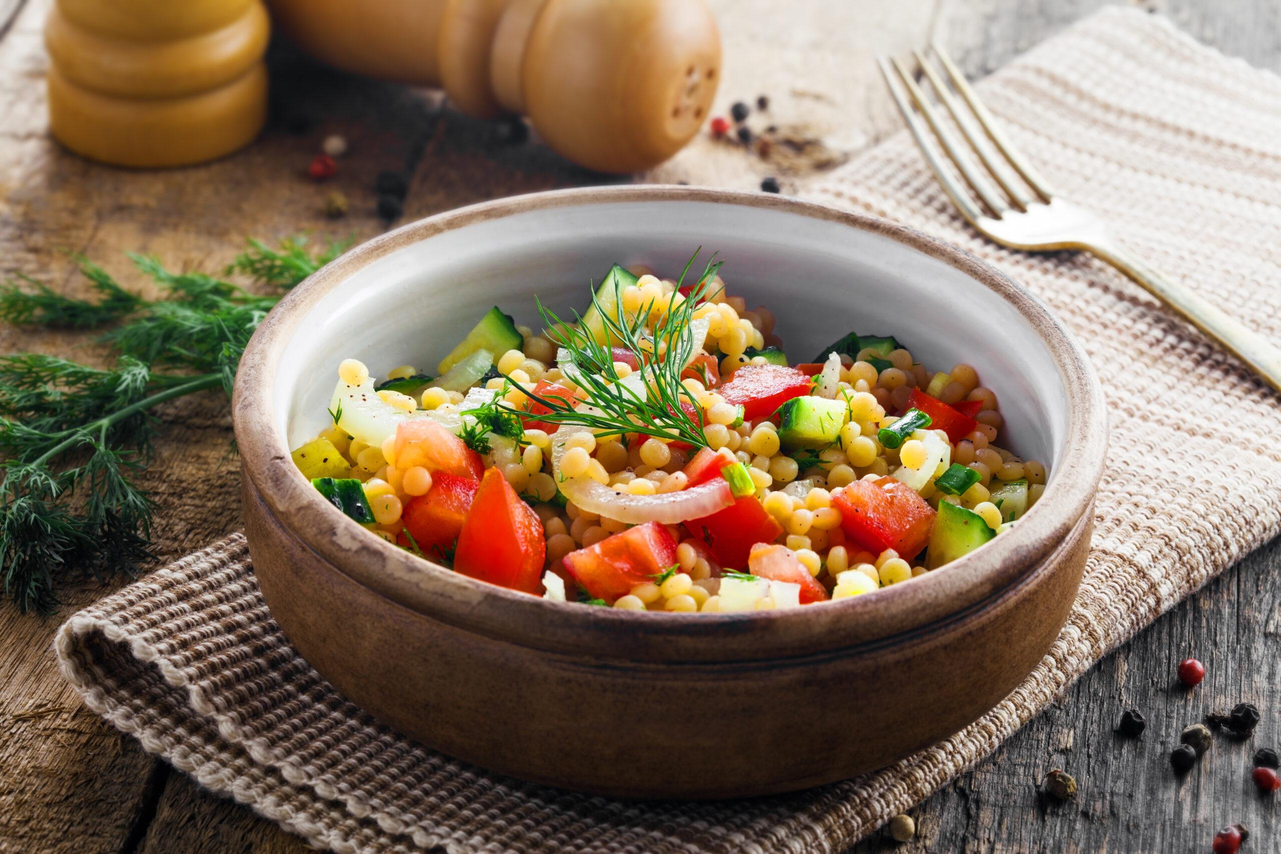Close-up of a bowl of couscous salad with chopped tomato, cucumber, onion, and fresh dill on a wooden table.
