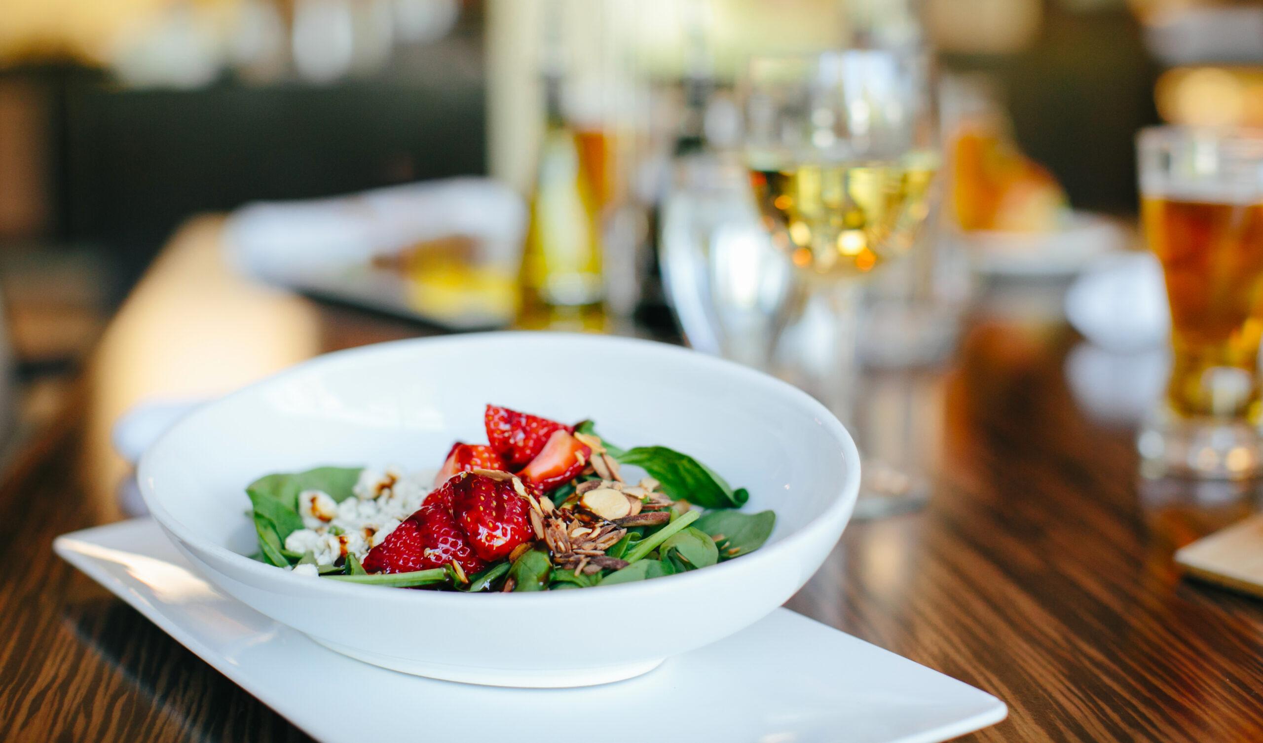 Fresh salad of spinach, sliced strawberries, feta cheese, and almond slivers served in a white bowl on a wooden table with beverages.