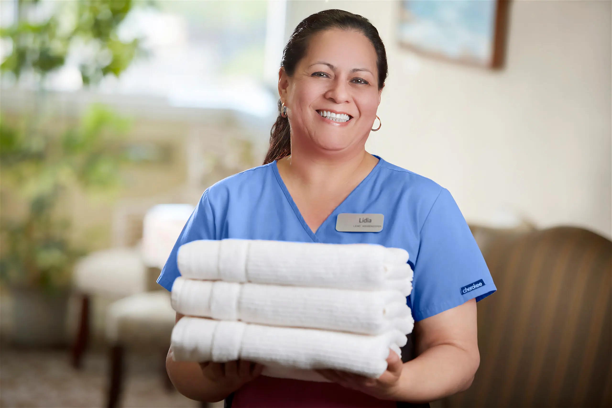 A housekeeping staff member in a blue scrub top and name badge smiling while holding a stack of folded white towels.