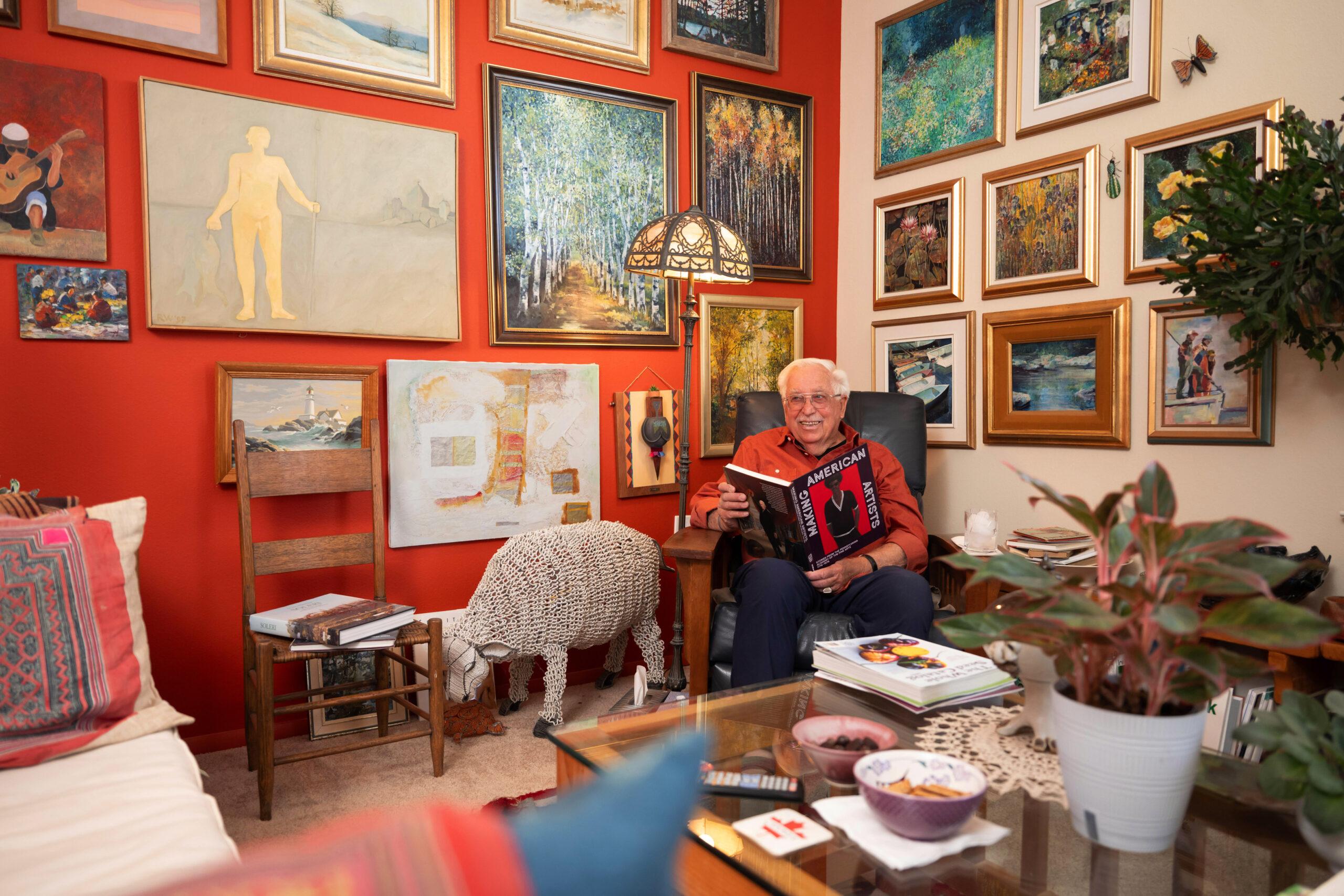 An older gentleman wearing glasses and a red shirt standing in a room filled with pottery and artwork on shelves.