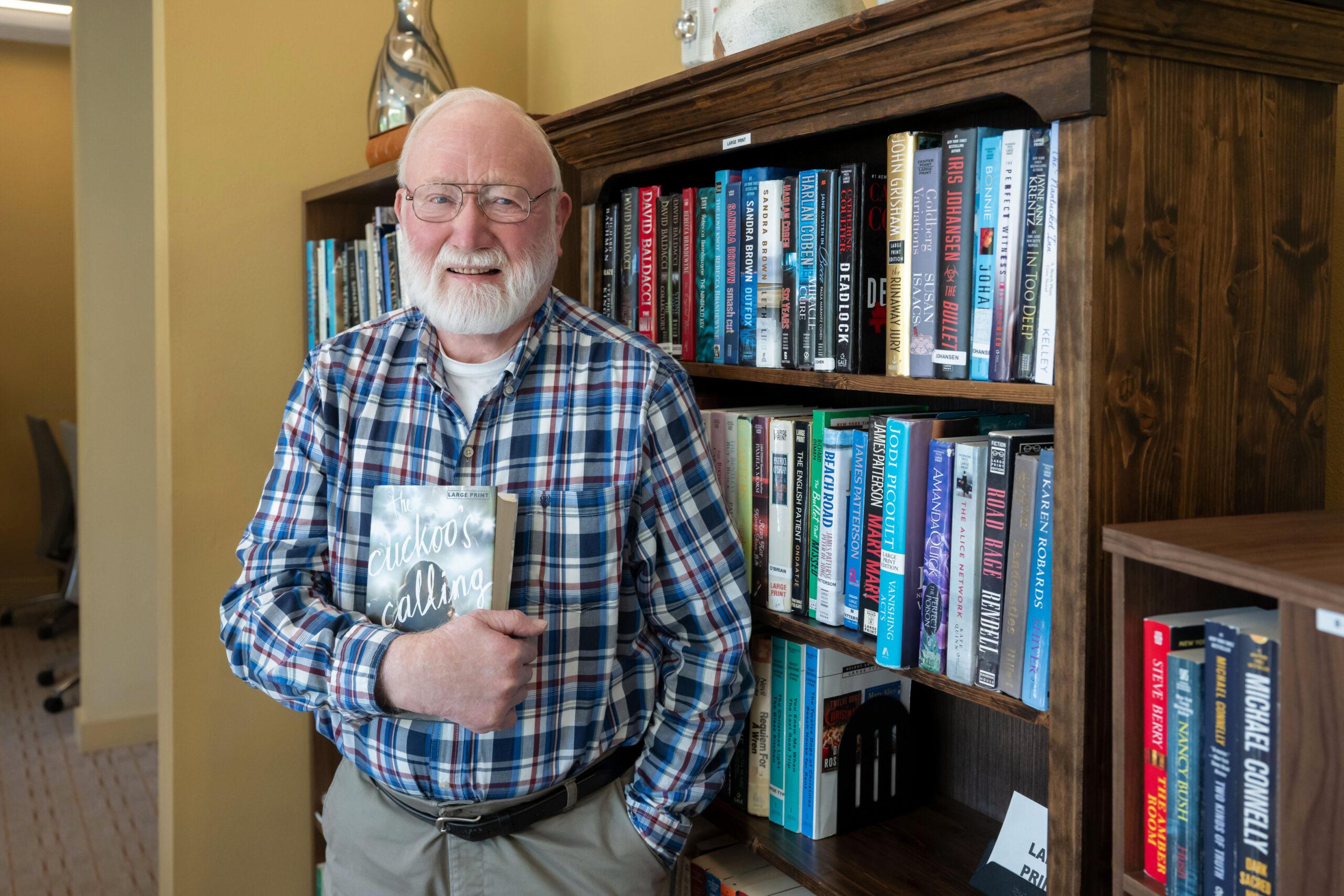 An elderly man wearing glasses and a plaid shirt holding a book titled 'The Cuckoo's Calling' in front of a bookshelf full of books.