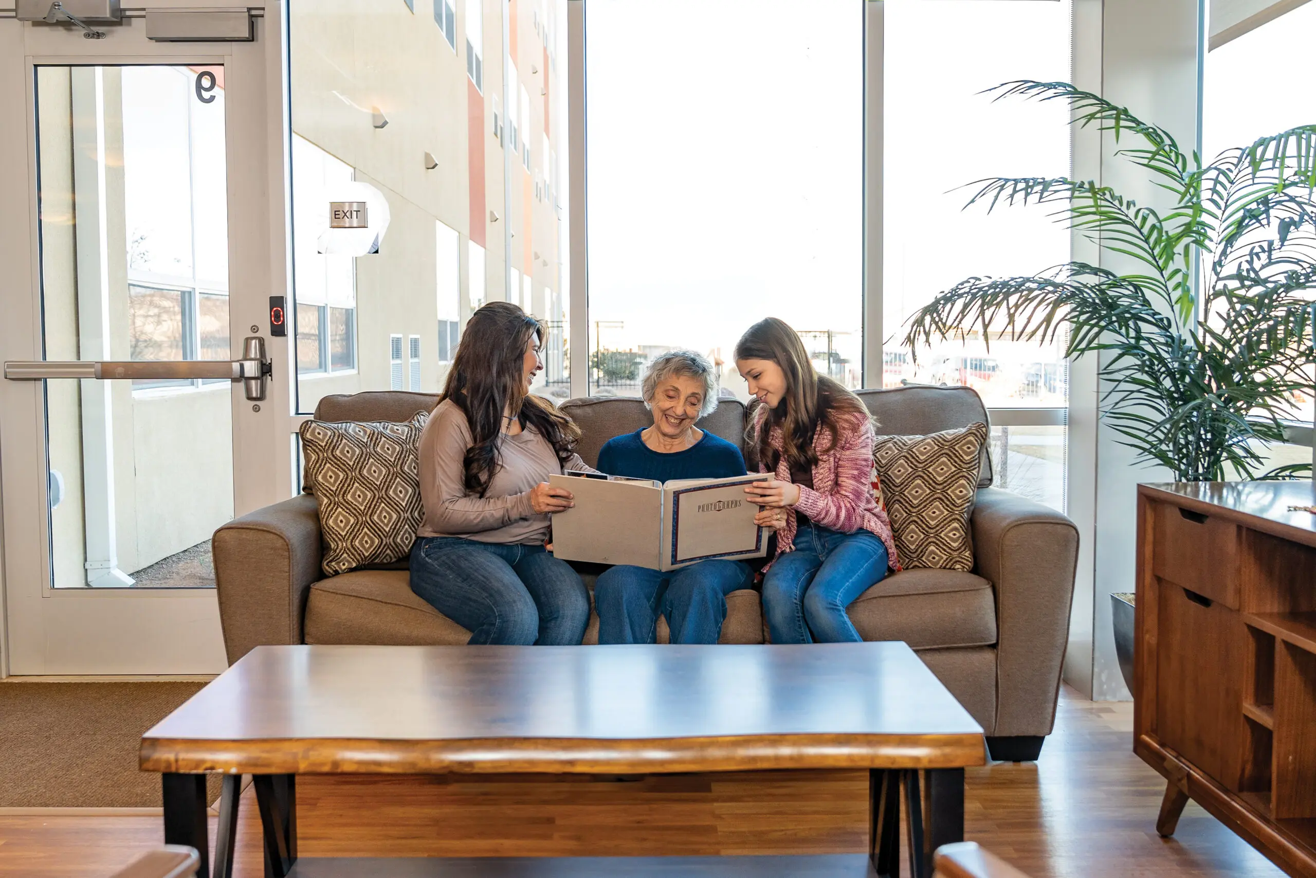 An elderly woman and two younger women sitting on a sofa, looking through a photo album together.