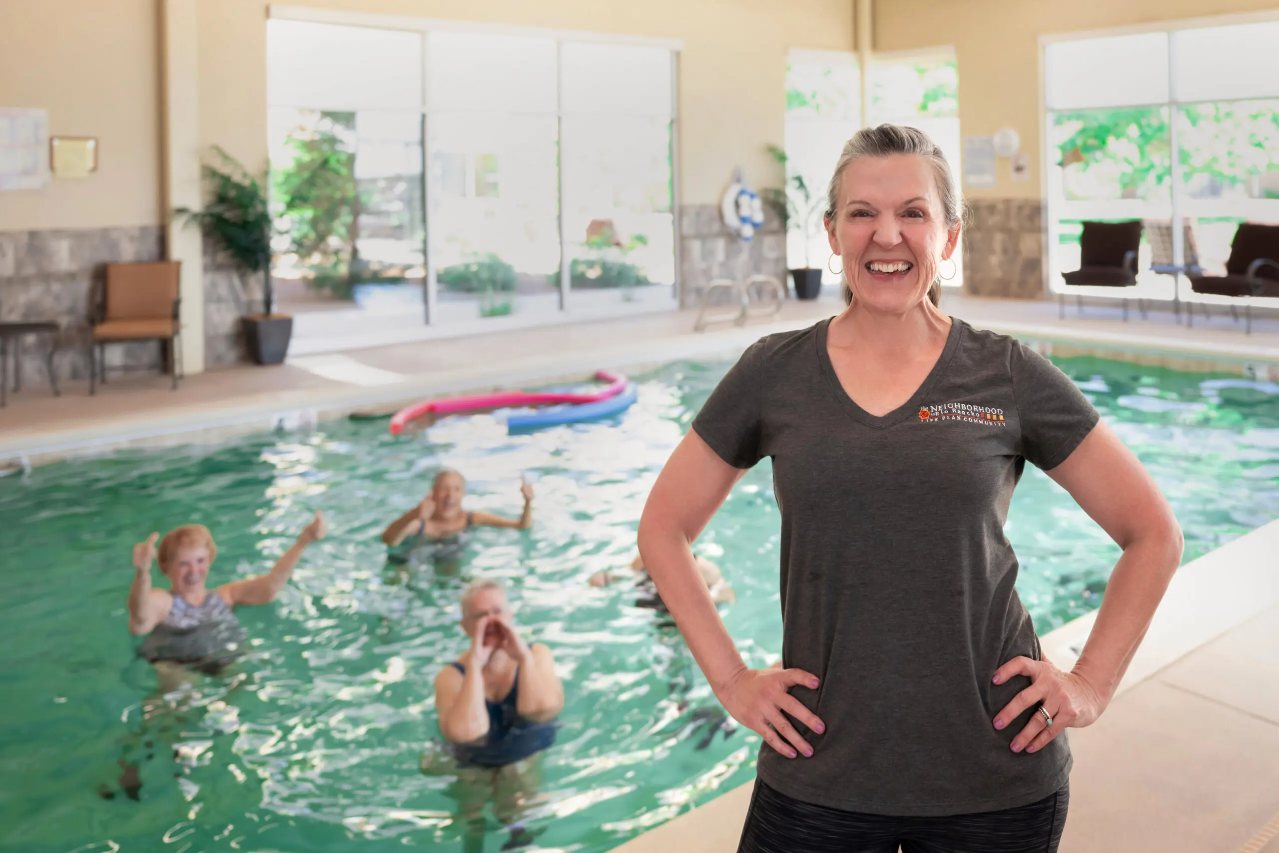A pool attendant in a gray t-shirt smiling in front of an indoor pool where seniors exercise in the water.