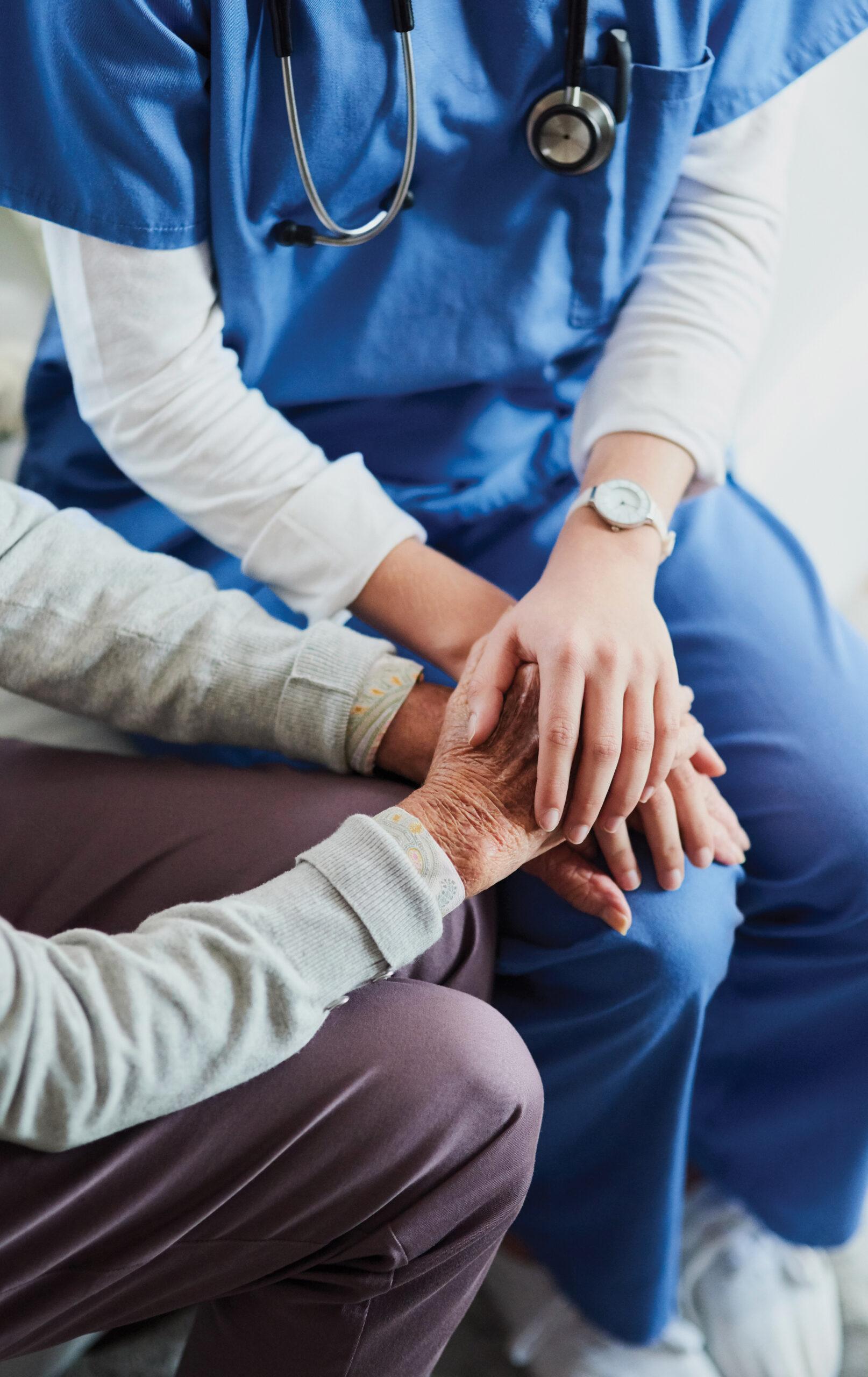 A nurse in blue scrubs gently holding the hand of an elderly patient.
