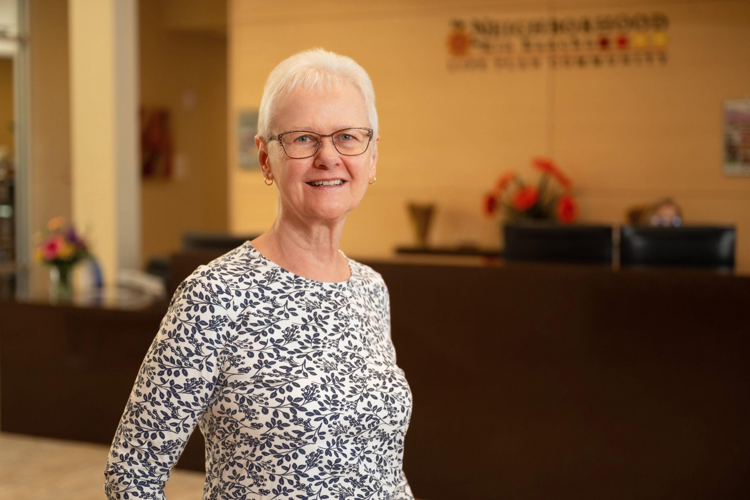 A smiling older woman in a patterned blouse standing in a community reception area with a sign on the wall behind her.