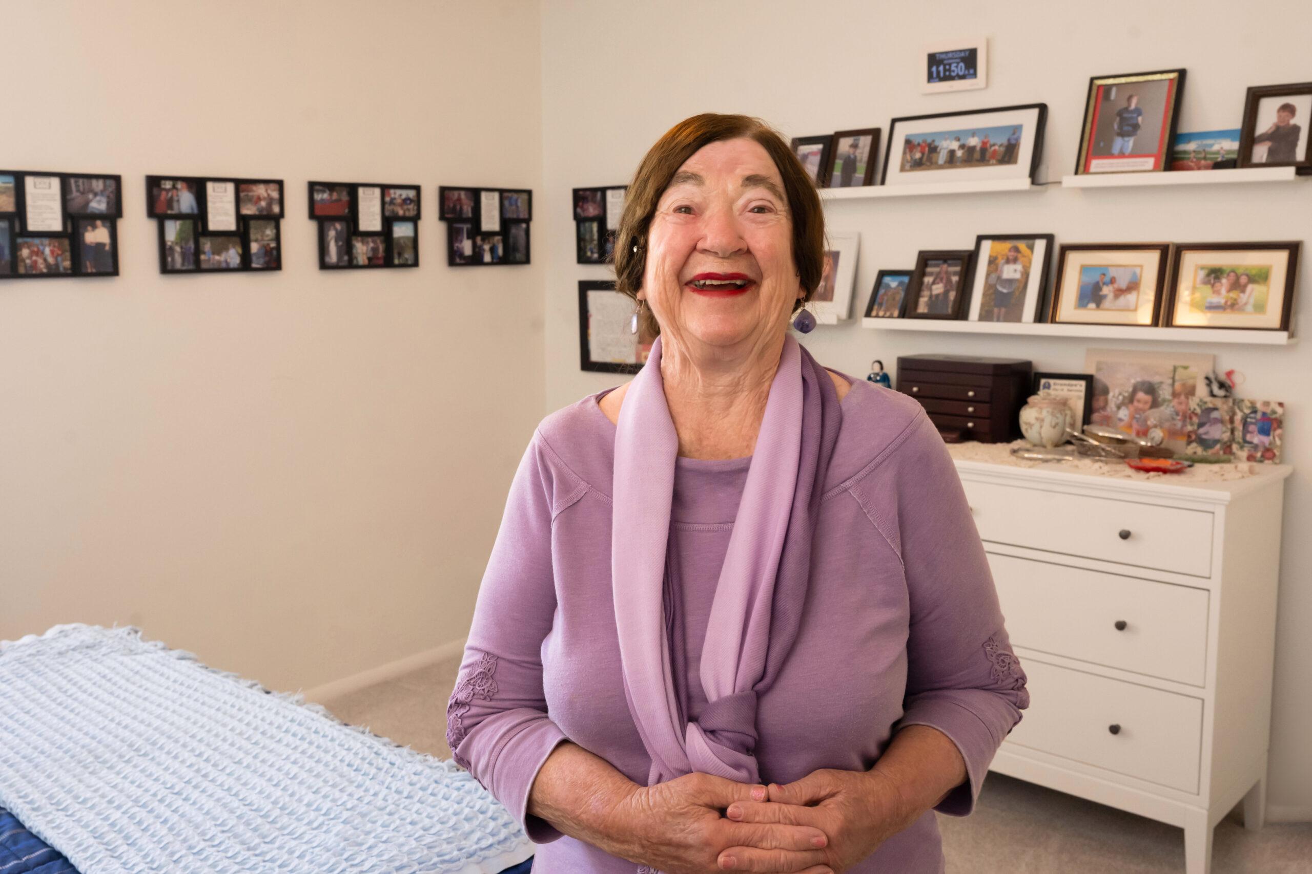 An older woman wearing a lavender top standing in a brightly lit room with family photos on shelves smiling at the camera.