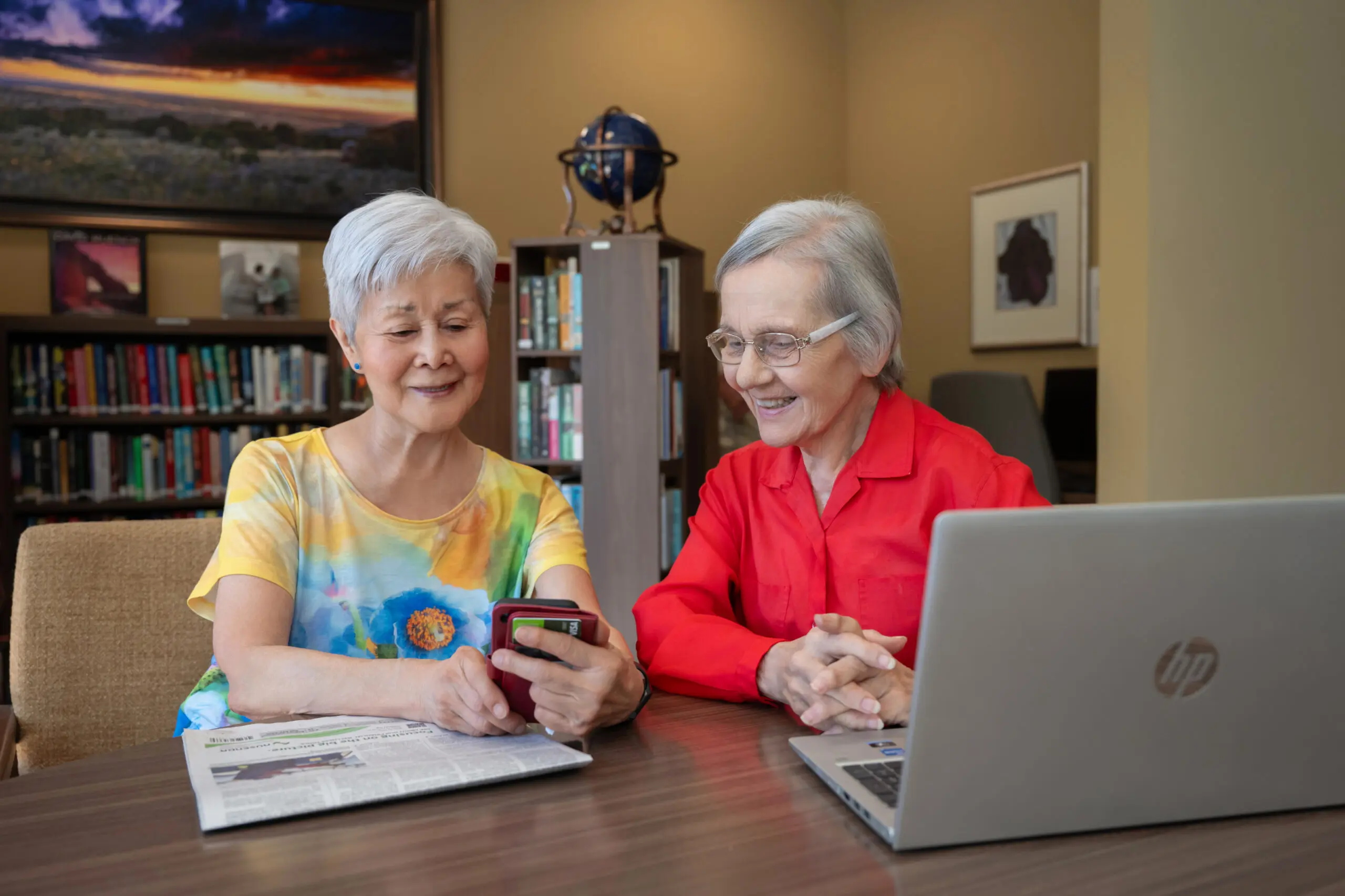 Two senior women sitting side by side at a table with a laptop and newspaper, one showing something on a phone to the other as they laugh.