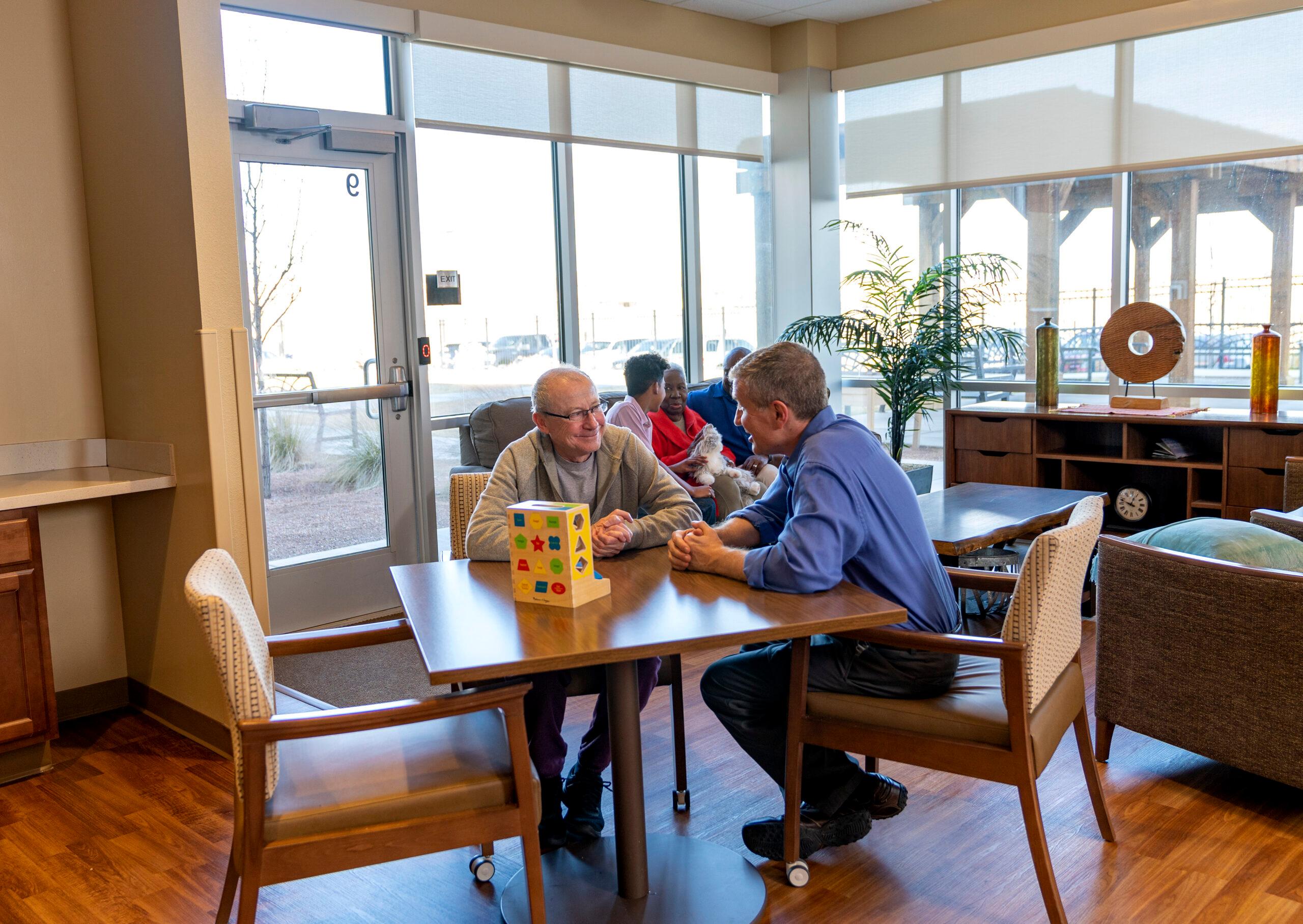 Memory care lounge with residents playing a tabletop activity near bright windows.
